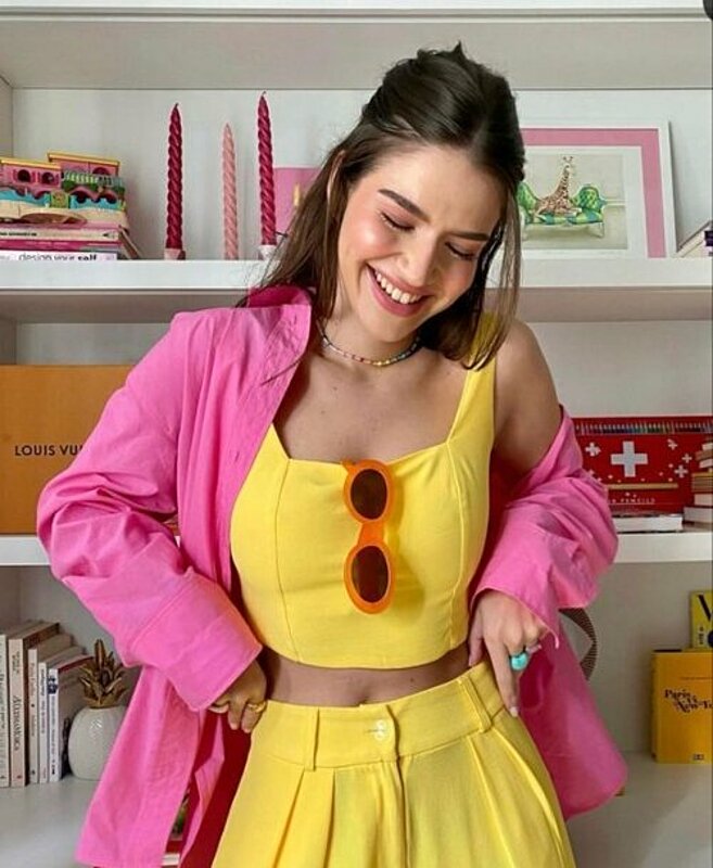 A woman wearing a yellow top and pink jacket, looking stylish and confident.